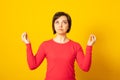 Peaceful woman meditates indoor, keeps hands in mudra gesture, tries to relax after long hours of work, holds fingers in yoga sign