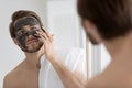 Peaceful young man applying black charcoal clay mask on face.