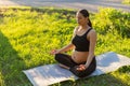 Peaceful young positive pregnant woman in gymnastic suit does yoga and meditate sitting on mat on green grass on sunny Royalty Free Stock Photo