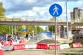 Pedestrian blue road sign and people walking on a sidewalk by a construction site, under a bridge.