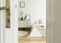 A peek through a half open door into a monochromatic beige and white bedroom interior with a bed and a drawer cabinet. Real photo.