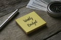 Pen, compass, banknotes and yellow sticky notes written with Weekly Budget on wooden background