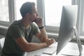 Pencive intelligent young man in casual t-shirt sits at the desk with trendy PC