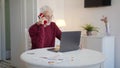 An elderly man sits at a table in front of a laptop and talks to a colleague on the phone