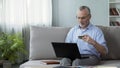 Pensioner sitting on sofa and inserting card number on laptop, online shopping