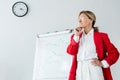 pensive attractive businesswoman in red jacket standing near flipchart