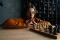 Pensive attractive young woman in elegant eyeglasses thinking about chess move sitting on floor in dark library room Royalty Free Stock Photo