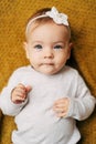 Pensive baby girl with a flower on her head in a white bodysuit lies on a yellow blanket, clenching her fists on her