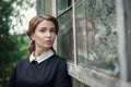 Pensive beautiful young girl in retro style dress standing near the window of old wooden house. Royalty Free Stock Photo