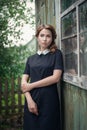 Pensive beautiful young girl in retro style dress standing near the window of old wooden house. Royalty Free Stock Photo