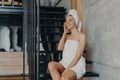 Pensive beautiful young woman applies body cream, enjoys softness of skin, sits on stairs at home, wrapped in white bath towel, Royalty Free Stock Photo