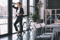 Pensive businesswoman in suit with digital tablet standing at window in office, bicycle standing behind