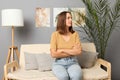 Pensive calm brown haired woman wearing beige T-shirt and jeans sitting on sofa in living room keeps hands folded looking away