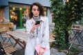 Pensive casual brunette girl in trench coat thoughtfully using cellphone on street