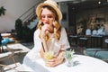 Pensive curly girl in vintage straw hat and white dress waiting boyfriend in cafe. Indoor portrait of sad fair-haired