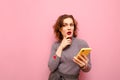 Pensive curly lady in gray clothes stands with smartphone in hand on pink background and looks away at copy space and thinks.