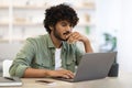 Pensive dark-skinned guy sitting at workdesk in front of laptop