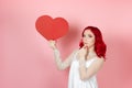 a pensive doubting hesitant woman in white dress and with red hair holds large paper red heart and rubs chin with hand