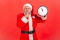 Pensive elderly man with gray beard wearing santa claus costume standing holding wall clock, holding Royalty Free Stock Photo