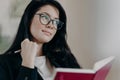 Pensive female owner of business works on planning schedule, writes in notepad, holds pen, looks thoughtfully away, wears formal