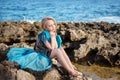 Pensive girl in azure dress with a tail like a bird sitting on a rocky seacoast of the ocean sea Royalty Free Stock Photo