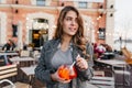 Pensive girl in elegant gray coat looking around, while waiting someone in outdoor cafe. Graceful curly woman with glass