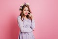 Pensive girl in flower wreath posing on pink background. Fascinating european lady in purple clothes standing in studio