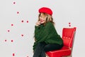 Pensive girl with shiny hair sitting on red chair and smiling. Good-looking french female model in knitted green sweater Royalty Free Stock Photo