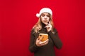 Pensive girl in sweater on hat of santa claus stands on red background with smartphone in hand, looks into screen and thinks. Royalty Free Stock Photo