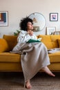 Pensive happy African american woman reading a book at home sitting on sofa relaxing drinking coffee. Vertical image. Royalty Free Stock Photo