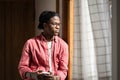 Pensive introverted African American man with mobile phone looking out window standing in house