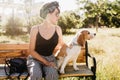 Pensive lady wears black ribbon in hair looking away while sitting on wooden bench with beagle dog. Outdoor portrait of