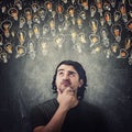 Pensive man, keeps hand under chin, thoughtful gesture, looking up suspicious at different light bulbs sketches. Puzzled and Royalty Free Stock Photo