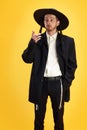 Pensive man in traditional Jewish attire with wide-brimmed hat and side curls posing against sunny yellow background.