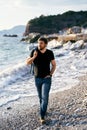 Pensive man walking on a pebble beach with a denim jacket over his shoulder against the background of the sea and rocks Royalty Free Stock Photo