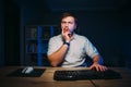 A pensive man works online on a computer at night in a bedroom with a blue light and looks intently at the screen. Bearded