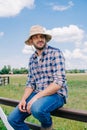 pensive middle aged farmer in panama hat sitting on fence and looking away