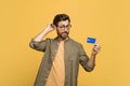 Pensive middle aged man scratching head, look at credit card and think over purchases, yellow studio background Royalty Free Stock Photo