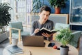 Pensive middle aged neutral gender woman making notes in paper notebook, using laptop, working in open space office Royalty Free Stock Photo