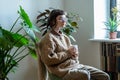 Pensive millennial girl sitting with phone and cup of tea in urban jungle home looking out window Royalty Free Stock Photo