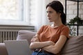 Pensive millennial lady sitting on couch using laptop planning work