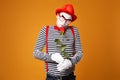 Pensive mime man with white face in red hat and striped t-shirt holding rose isolated on empty orange background Royalty Free Stock Photo