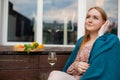 Pensive, mindful, calm blond woman in pink shirt, covered by blue blanket with wineglass and plate of food on backyard