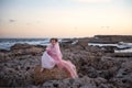A pensive, offended princess in a pink dress with a veil and crown sitting cringing on a rock and the sky glow from dawn sunrise Royalty Free Stock Photo
