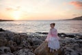 Pensive sad princess girl in a pink dress and a diadem is sitting on a rock on the ocean seashore and meets the dawn of the sunris