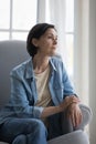 Pensive serious middle-aged woman sits on armchair indoors