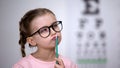 Pensive smart girl wearing glasses, testing vision with eye chart in clinic