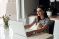 Pensive, smiling young brown long haired woman sitting in white kitchen, use laptop. Freelancer and self-employed worker Royalty Free Stock Photo