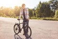 Pensive teenage boy standing on road, keeping hand on handle bar of his bike, waiting for others cyclists to have journey together