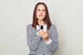 Pensive thoughtful brown haired adult woman wearing striped shirt standing isolated over gray background using mobile phone Royalty Free Stock Photo